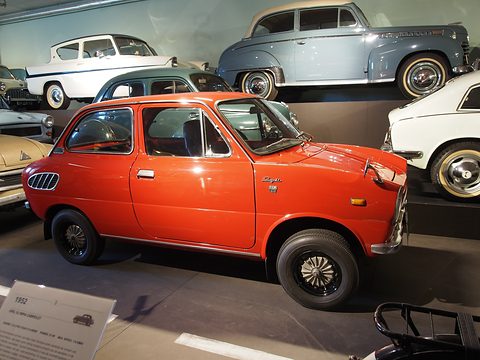 Orange small saloon with whitewall tyres on a dark museum floor, other old cars on a shelf behind. Opens a larger view