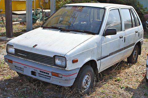 White small five-door car parked on dry grass and straw, a yellow sign behind. Opens a larger view