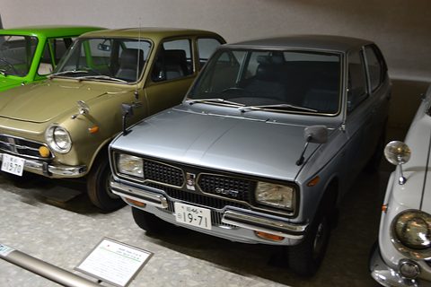 Grey small saloon with square headlamps in a museum row, a placard on the floor. Opens a larger view