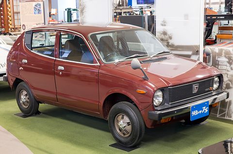 Maroon small car with a black grille and a blue plate on green carpet, displays behind. Opens a larger view