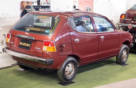 Rear three-quarter of a maroon small car with SUZUKI on its tail panel, on green carpet. Opens a larger view