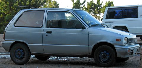 Side view of a pale blue small five-door car on a muddy lot, a white van behind. Opens a larger view