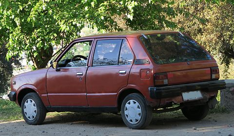 Rear three-quarter of a faded red five-door car parked under trees. Opens a larger view