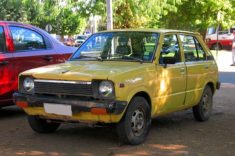 Yellow small five-door car parked under trees beside a red car. Opens a larger view