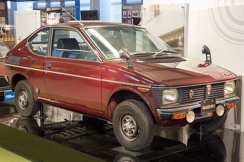 Low maroon two-door fastback with four round headlamps, wing mirrors and a chrome bumper, on a black museum plinth. Opens a larger view