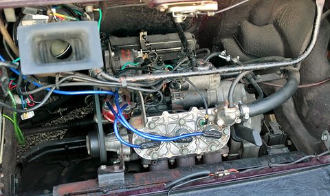Engine bay with a small three-cylinder engine, an alloy cover, blue ignition leads and an air intake box above.