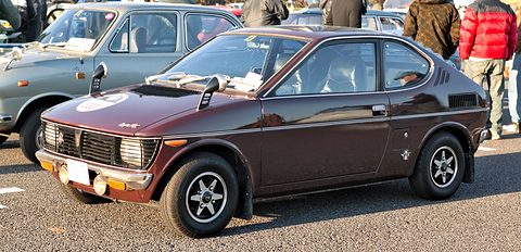Low maroon two-door fastback with four round headlamps in a black grille, wing mirrors and alloy wheels, on a car park with visitors behind. Opens a larger view
