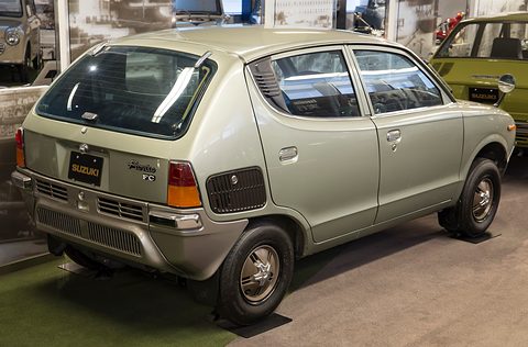 Rear three-quarter of a pale gold four-door small car on a dark floor with photographs behind. Opens a larger view