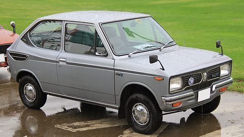 Silver small saloon on wet tarmac beside bright green grass, rain drops on its roof. Opens a larger view