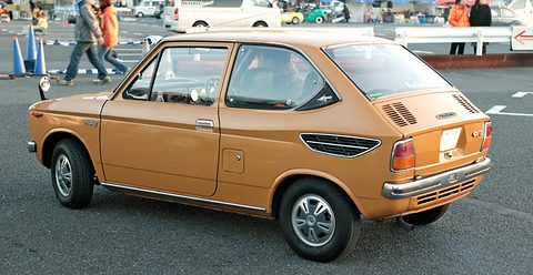 Side view of an orange small hatchback-shaped saloon on tarmac, people walking behind. Opens a larger view