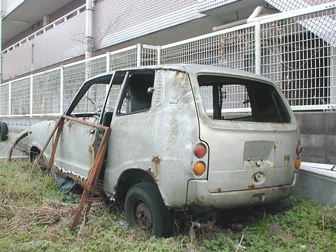 A rusty, doorless small van-like car in long grass against a white fence. Opens a larger view