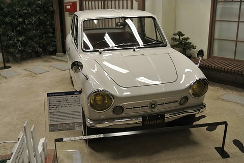 White small saloon seen from the front, yellow headlamps and an information board beside it. Opens a larger view
