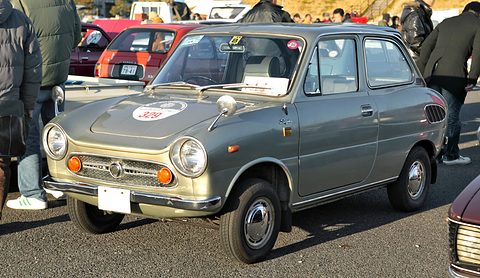 Grey small saloon with round headlamps and amber fog lamps, visitors in winter coats standing around it. Opens a larger view