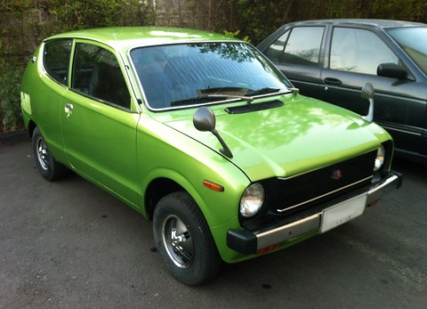 Lime-green small car with a black grille, parked on a street in the shade of trees. Opens a larger view