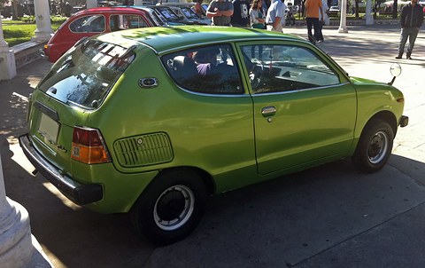 Rear three-quarter of a lime-green small car with a ribbed side vent, in bright sun on a plaza. Opens a larger view