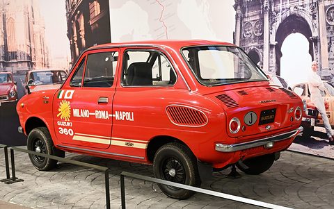Rear three-quarter of a red saloon lettered MILANO–ROMA–NAPOLI, a map of Italy on the wall behind. Opens a larger view