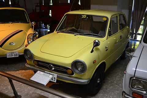 Yellow small saloon in a museum row, an orange car with black stripes to its left. Opens a larger view