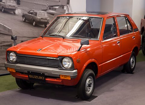 Orange-red small five-door car on a grey floor in front of a black-and-white photo wall. Opens a larger view