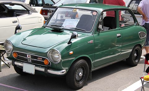 Dark green small saloon with round headlamps and amber lamps, parked beside a camping chair. Opens a larger view