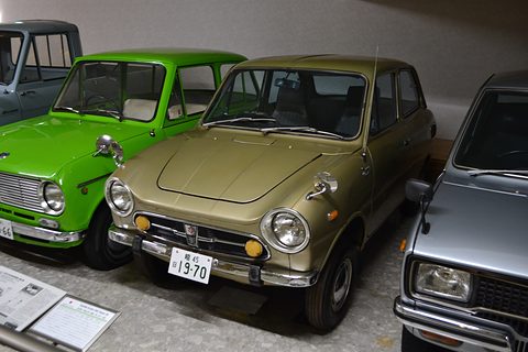 Olive-gold small saloon between a lime-green car and a grey car in a museum row. Opens a larger view