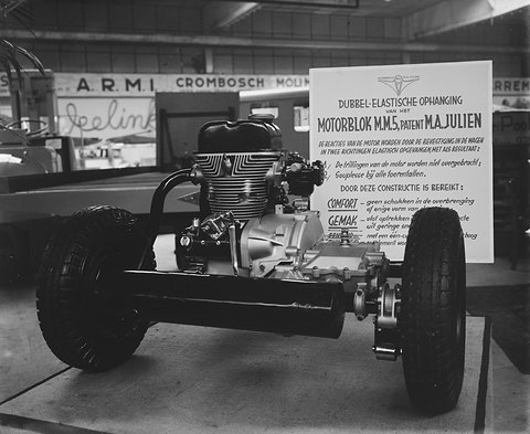 Black-and-white: a bare chassis with a finned single-cylinder engine and fat tyres in front of a lettered sign.