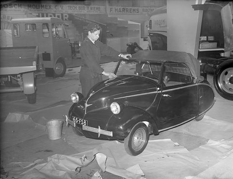 Black-and-white: a dark rounded two-seater with a folding hood being wiped by a woman in overalls, vans behind.