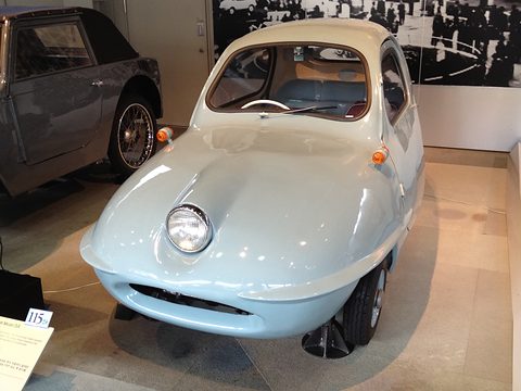 Pale blue rounded three-wheeler from slightly above, showing its one headlamp, curved windscreen and seats inside. Opens a larger view