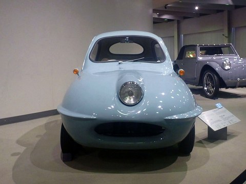 Front view of a rounded pale blue three-wheeler with one central headlamp above a wide oval opening. Opens a larger view