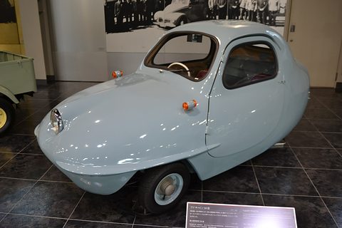 Pale blue-grey teardrop three-wheeler with a single headlamp and orange indicators on a dark tiled floor, a label board in front.