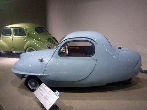 Side view of a pale blue three-wheeled car with a long tapering tail, one door and a museum label, a green saloon behind.