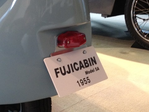 White plate with black lettering under a small red tail lamp on a pale blue rounded body. Opens a larger view
