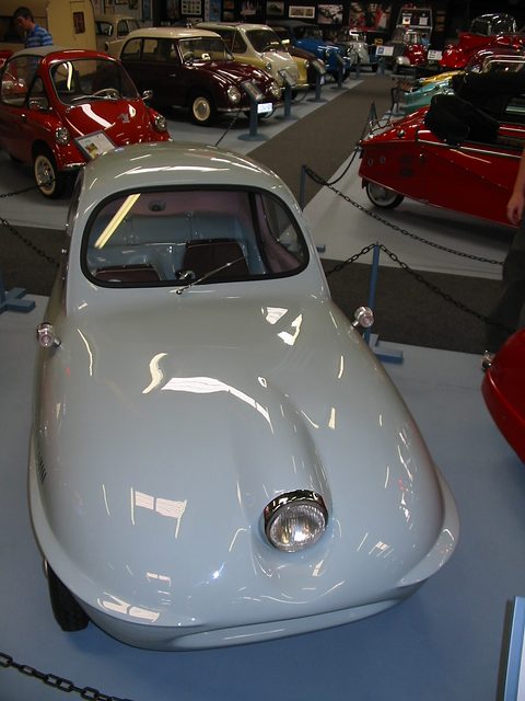 Silver rounded three-wheeler seen from the front and above, with rows of Isettas and other small cars behind. Opens a larger view