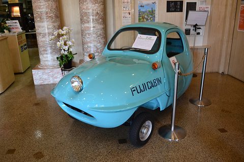 Turquoise three-wheeled bubble car with FUJICABIN on its side standing behind rope barriers in a lobby with marble pillars. Opens a larger view