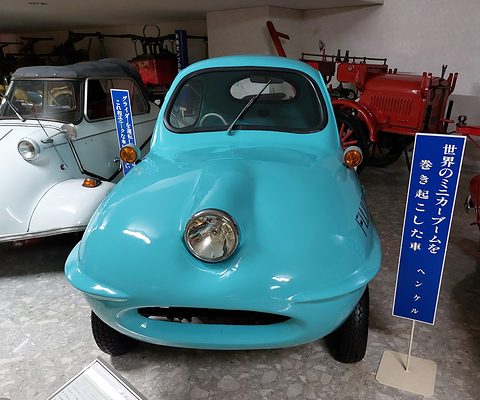 Rounded turquoise three-wheeler with a single central headlamp and a wide slot under its nose, beside a blue sign. Opens a larger view