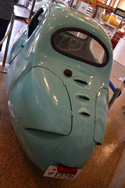 Rounded turquoise rear of a small car with an oval rear window, three slots above the engine lid and a red tail lamp. Opens a larger view