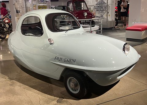 Very pale green rounded three-wheeler with a long nose, one headlamp and black lettering on its flank, other cars behind.