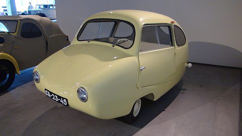 Cream egg-shaped three-wheeler on a grey floor beside a beige vintage car. Opens a larger view