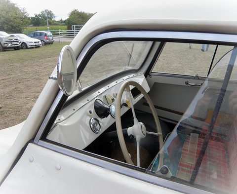 Through an open window: cream steering wheel, simple dashboard and tartan seat of a small car. Opens a larger view
