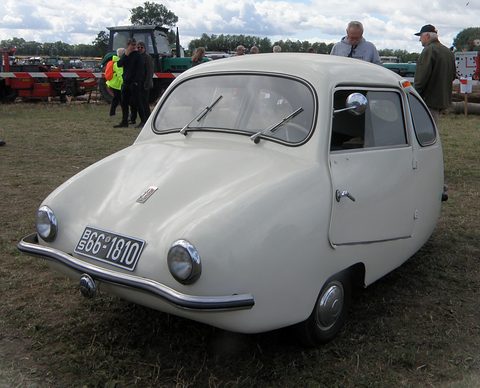 White rounded car with chrome bumper and German plate on grass, tractors and visitors behind. Opens a larger view