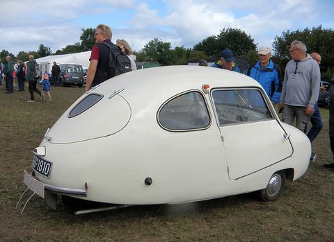White egg-shaped three-wheeler on a grass field, visitors gathered round it. Opens a larger view
