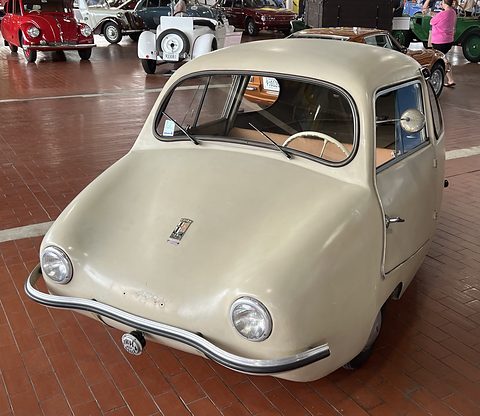 Cream rounded three-wheeler with a chrome bumper on a brick-red floor, rows of classic cars behind. Opens a larger view
