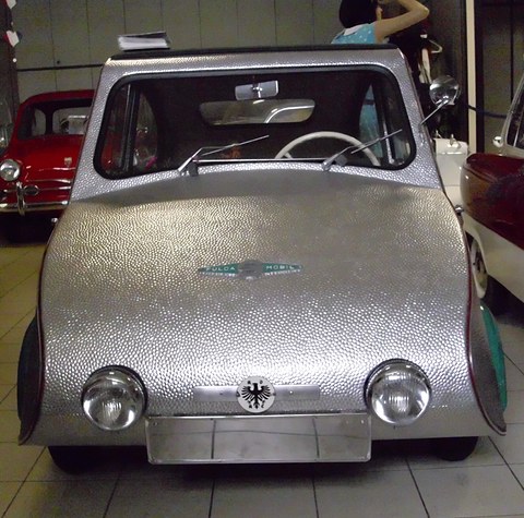 Silver three-wheeler facing the camera, hammered metal nose with two headlamps and a blank plate. Opens a larger view