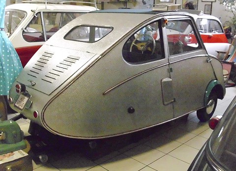 Dimpled aluminium three-wheeler seen from behind and the side among other classic cars on a tiled floor. Opens a larger view