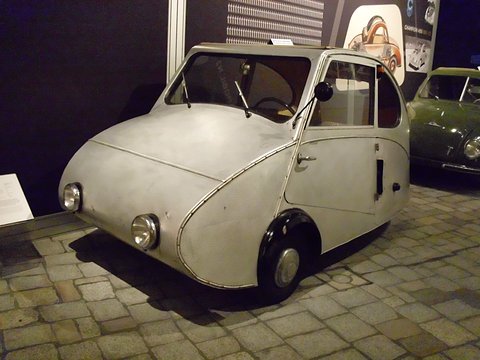 Pale silver three-wheeler with a pointed nose and two headlamps on a cobbled display floor, a dark wall behind. Opens a larger view