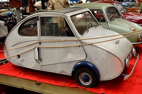 Hammered-aluminium three-wheeler with blue wheel arches on a red platform, a display rope across its flank. Opens a larger view