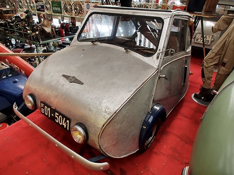 Dimpled silver three-wheeler with a front bumper bar and a German plate on a red platform among old vehicles. Opens a larger view