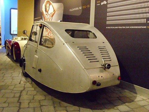 Silver three-wheeler from behind: louvred rear panel, small rear window and a single rear wheel, beside a display panel. Opens a larger view