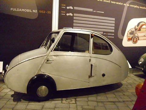 Silver three-wheeler in side view, its curved tail and front door clear, in front of a dark wall of graphics. Opens a larger view