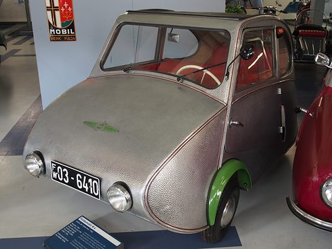 Hammered-aluminium three-wheeler with green wheel arches beside a Fulda Mobil enamel sign. Opens a larger view