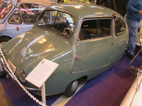 Grey-green rounded car with a front-hinged door on a purple carpet, an information board and chain barrier beside it. Opens a larger view